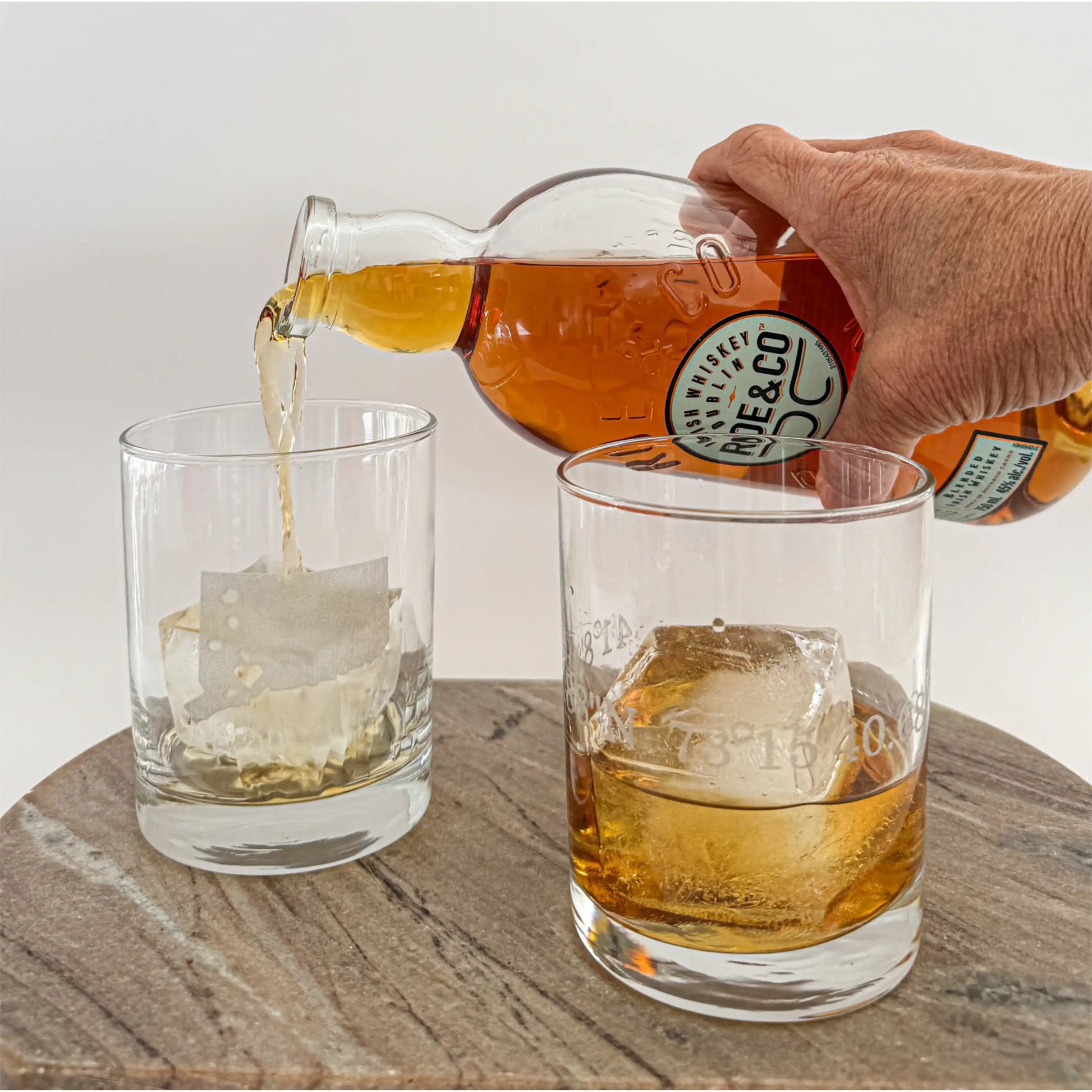 Person pouring a drink from a bottle labeled 'ROE&CO' into two glasses on a wooden surface.