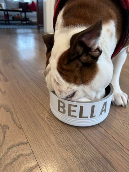 Dog eating from a white bowl labeled 'Bella' on a wooden floor.
