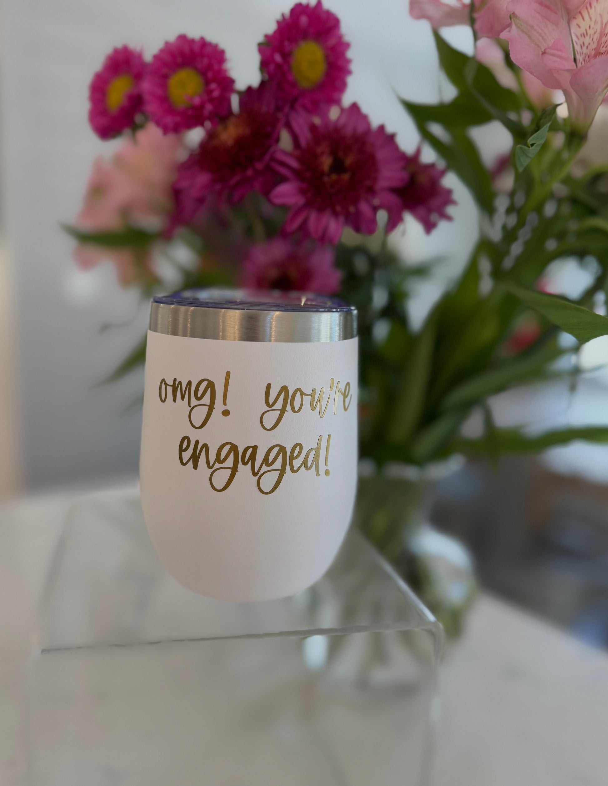 A white, stemless stainless steel tumbler with gold text that reads 'OMG! You're engaged!' in front of a bouquet of flowers.