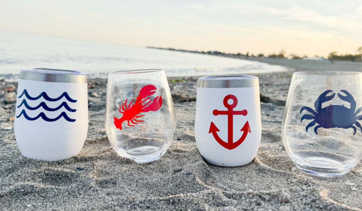 Four stemless wine glasses with beach-themed designs on a sandy surface.